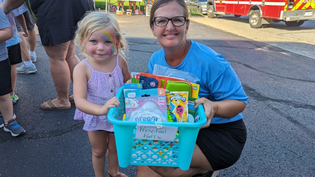 woman in blue shirt smiling handing basket of school supplies to small child